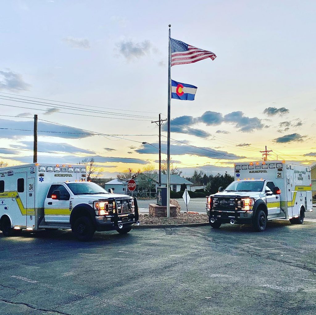 Two ambulances parked next to a flagpole with the American and Colorado flags